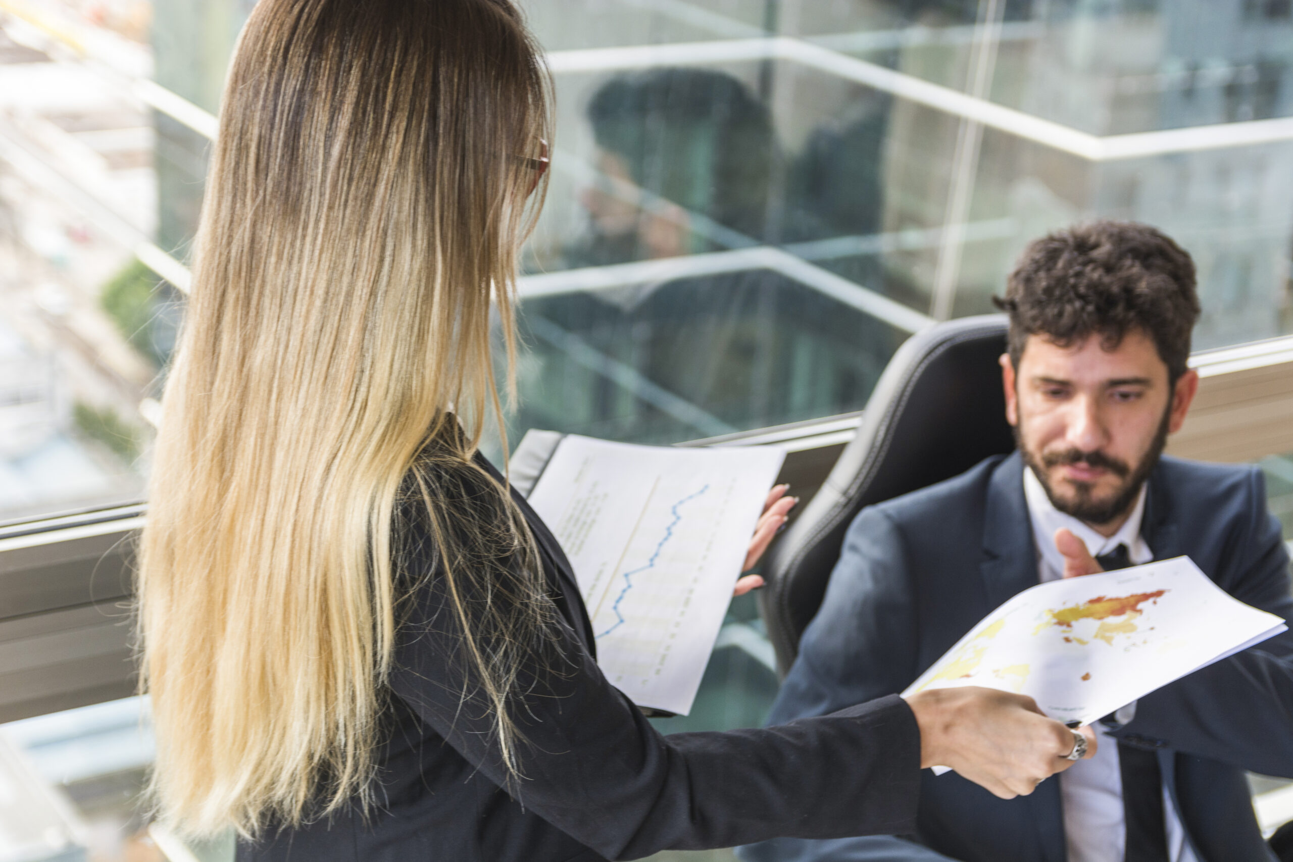 Zbliżenie w biurze: kobieta o długich blond włosach, ubrana w czarną marynarkę, podaje mężczyźnie w garniturze dokument z kolorową mapą świata. Mężczyzna siedzi w czarnym fotelu biurowym i z uwagą patrzy na przekazywane papiery. W tle widać duże okno z widokiem na miasto i odbicia w szybie.