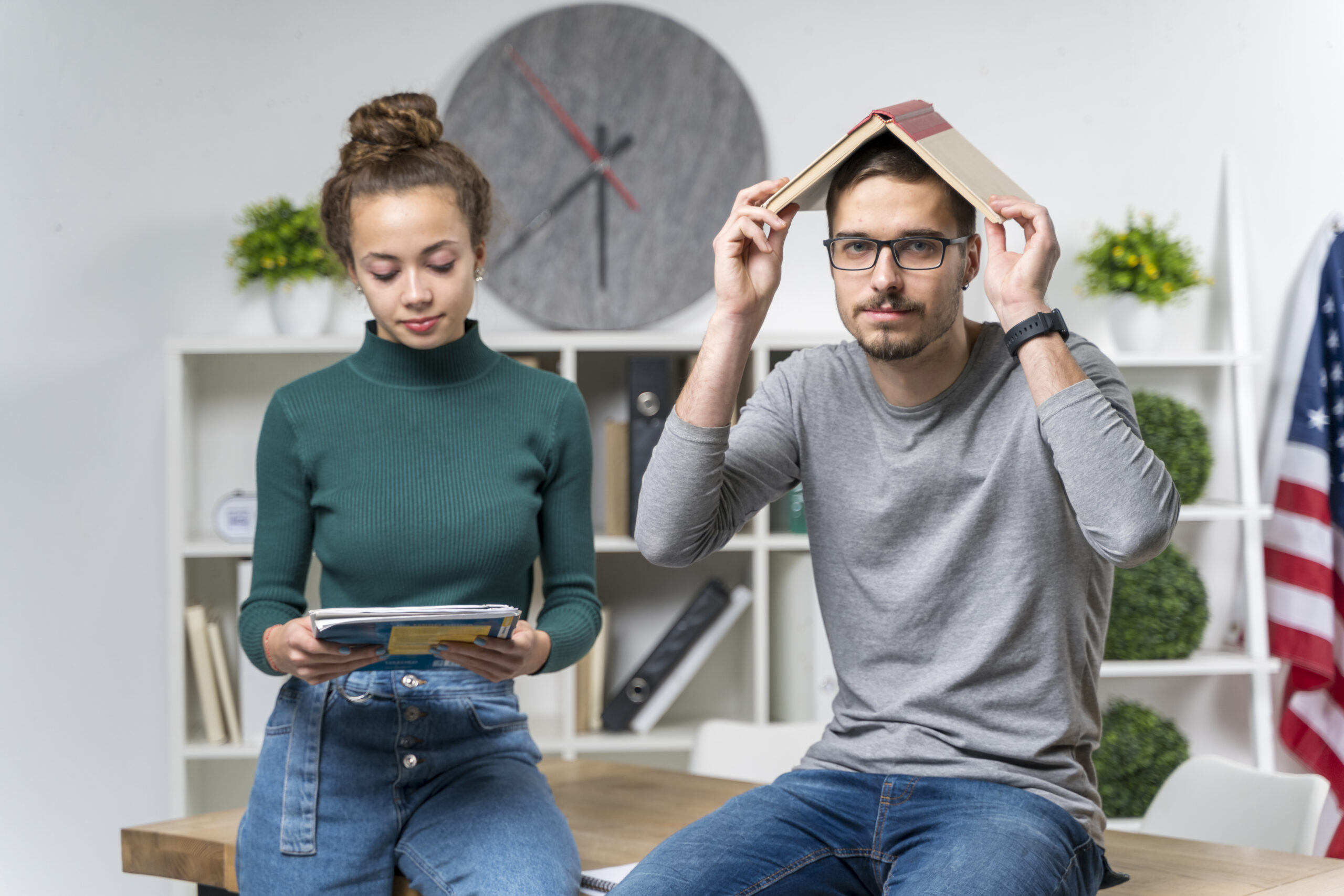 Zdjęcie dwojga młodych ludzi w biurze. Mężczyzna w okularach i szarej bluzie siedzi na biurku z otwartą książką na głowie, tworzącą kształt daszka. Obok niego stoi kobieta w zielonym golfie i dżinsach, patrząca na trzymany w rękach tablet. W tle widoczne regały z segregatorami, rośliny doniczkowe, duży zegar ścienny oraz flaga USA.