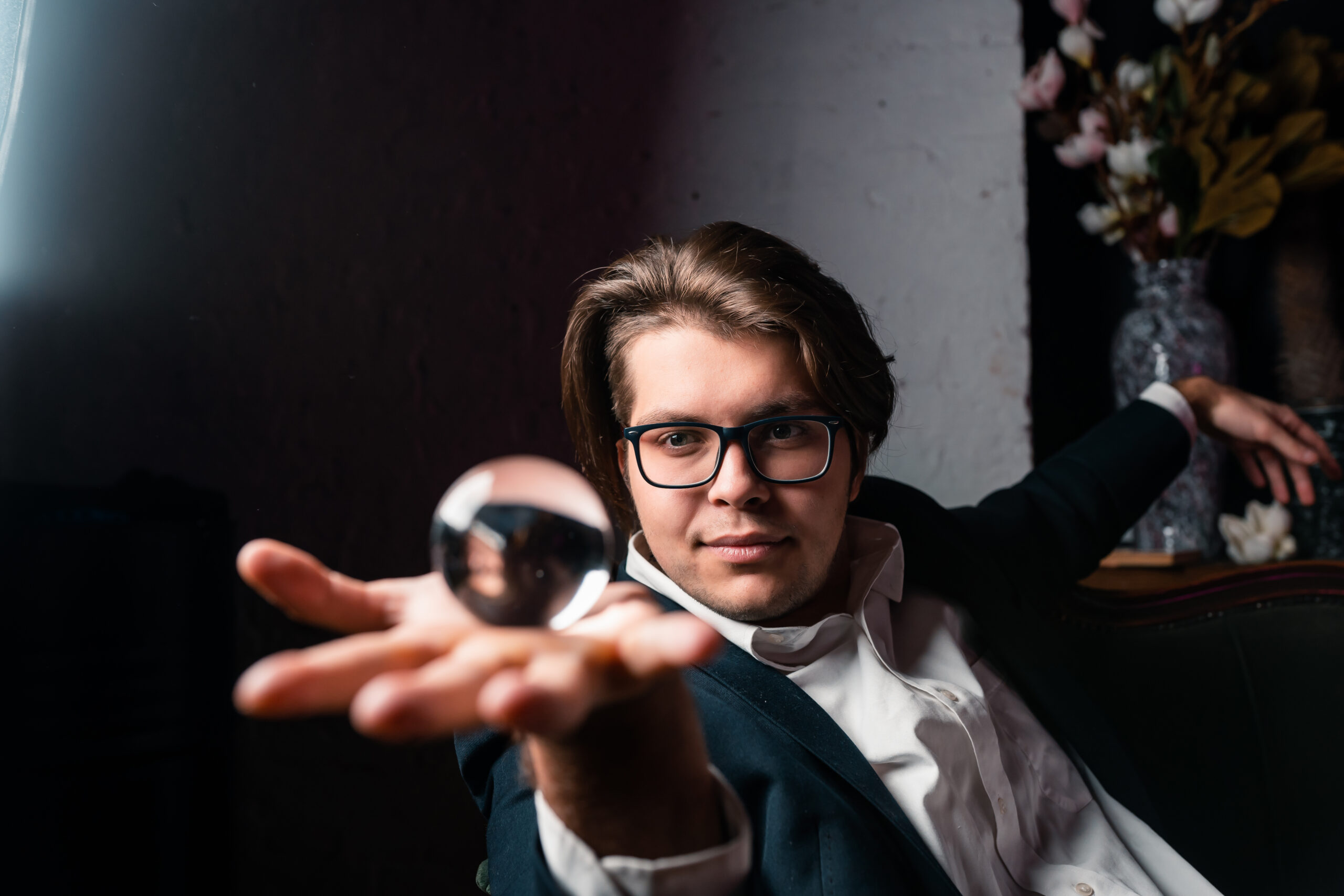 Młody mężczyzna w garniturze i okularach wykonujący trik magiczny z przezroczystą szklaną kulą - idealny iluzjonista na event w Warszawie.